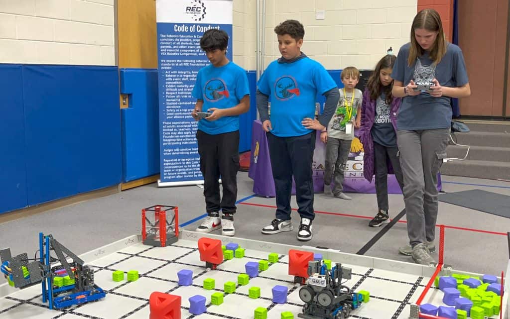 Two male 5th grade students from Indian Peaks standing side by side in front of a robotics field. The boy on the left is holding a controlling and moving his robot around the field. A taller female student from another school is to the right of them and also controlling her robot with a controller.