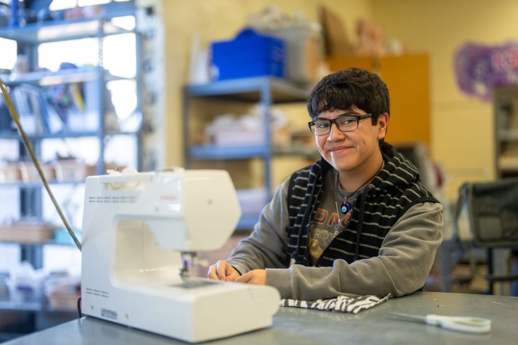 A student smiles while seated at a sewing machine, surrounded by crafting materials in a classroom setting.