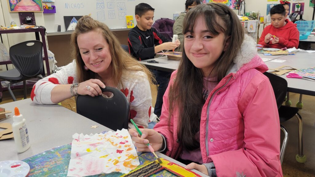 Artist and guest art teacher, Joyanna Rose Gittings, squatting down at a table next to a 5th grade girl with long hair who is sitting at a table and holding a paintbrush while she paints a Mexican nicho.