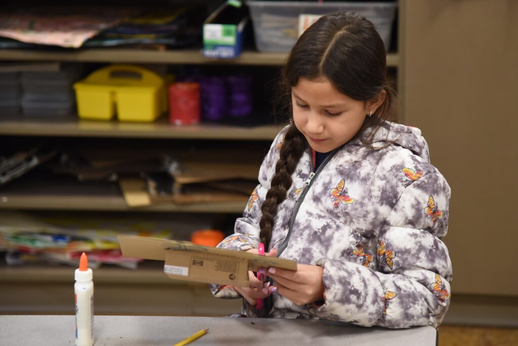 5th grade girl with long hair in a braid, sitting at a table looking down as she cuts a piece of cardboard into a nicho, a type of Mexican shadow box and photo frame.