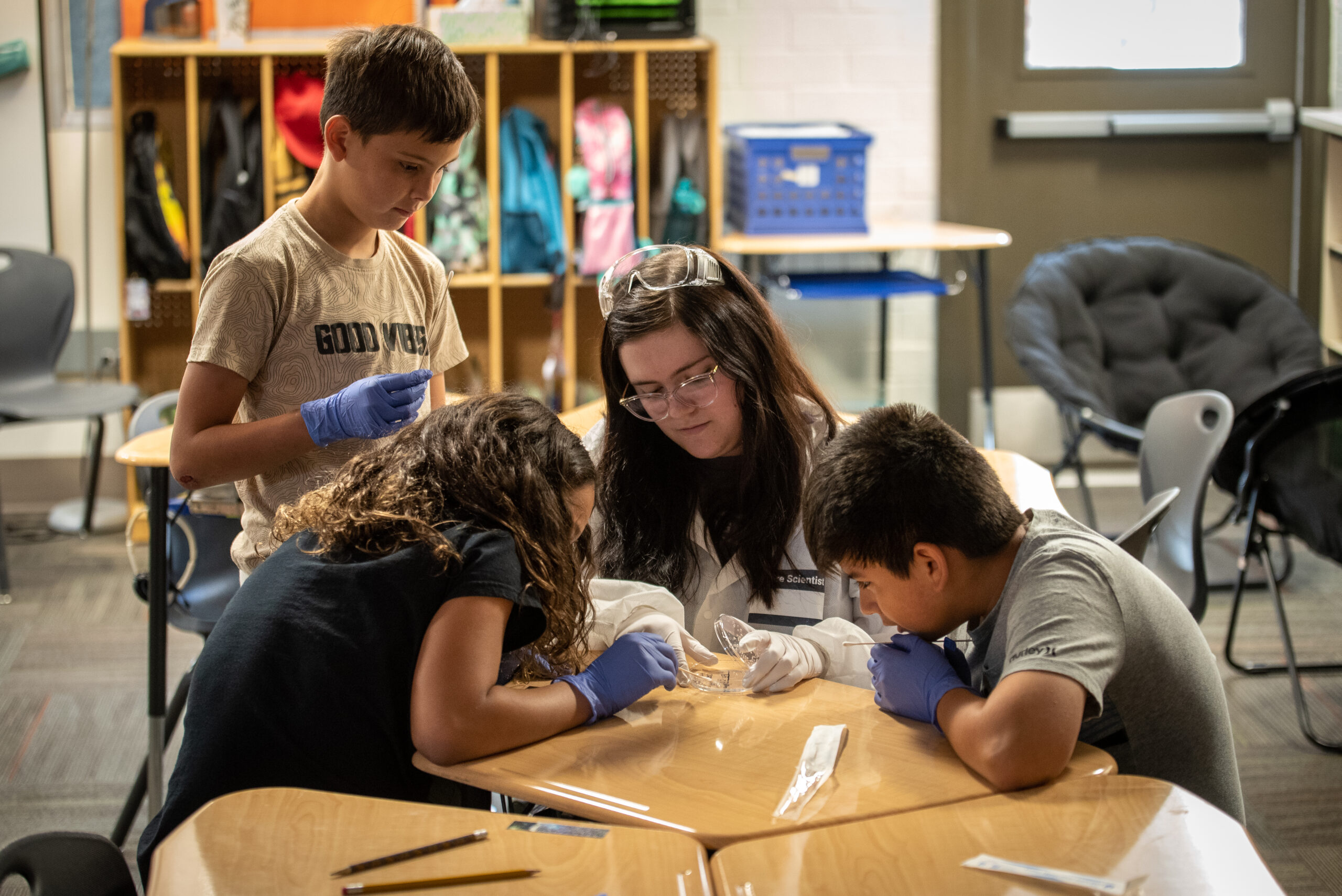 Estudiantes de primaria que rodean un preparatoria estudiante mientras miraba fijamente una placa de Petri sobre una mesa.