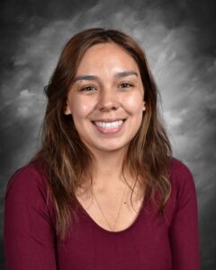 Portrait of a smiling woman with long brown hair, wearing a maroon shirt, against a gray background.